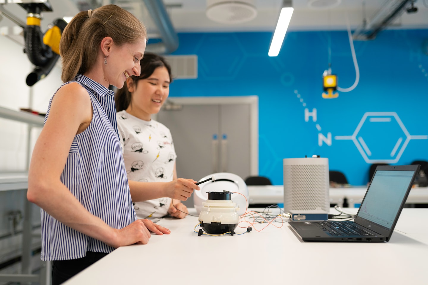 Two women examine a scientific device and laptop in a lab.